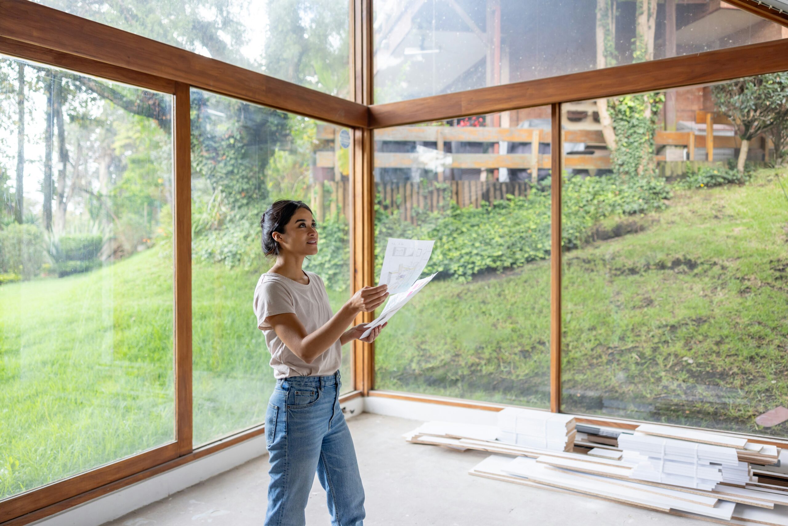 A woman reviews blueprints, modern room with large windows, signifying the start of a home remodeling project.